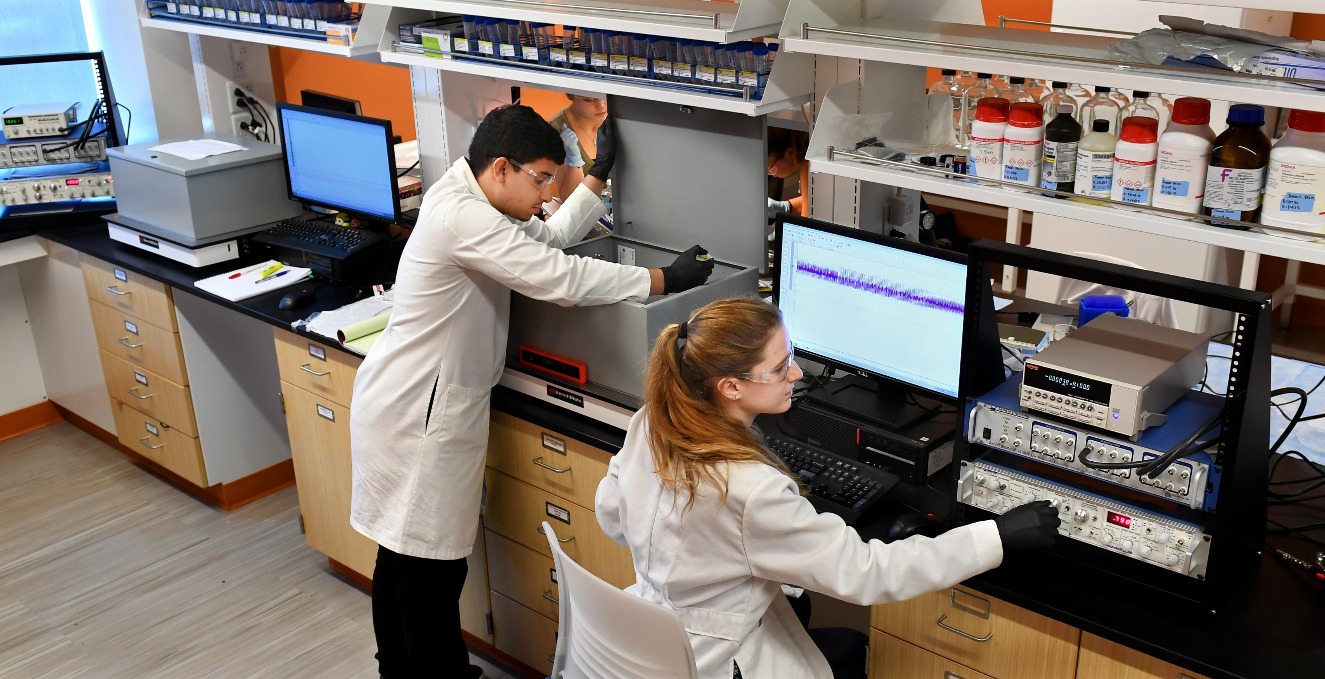 one researcher places something in a case, while another researcher reviews information on a computer in a lab